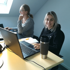 Zwei Frauen sitzen an einem Schreibtisch vor ihren Laptops. Die vordere Person lächelt in die Kamera.